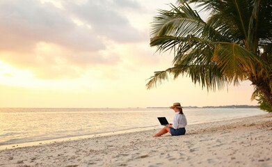 Young female freelancer wearing straw hat working on laptop while sitting on tropical beach at sunset