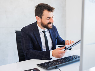 Handsome businessman in suit sitting in office by the computer, looking at documents and smiling.