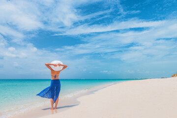 Beach vacation bikini woman enjoying summer travel Caribbean holiday with arms behind head relaxing in the wind at blue ocean walking with sun hat and pareo skirt.