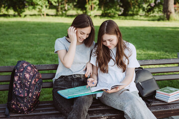 Wall Mural - Two student girls are looking at an open book on a bench in the park. Distance education, preparation for exams. Soft selective focus.