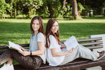 Wall Mural - Two student girls are looking at an open book on a bench in the park. Distance education, preparation for exams. Soft selective focus.