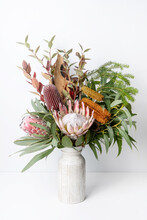 Beautiful Floral Arrangement Of Mostly Australian Native Flowers, Including King Protea, Banksia, Eucalyptus Leaves, Blue Berry Foliage And Rustic Bark, In A White Vase With A White Background.