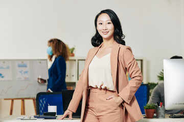 Sticker - Portrait of young confident beautiful female entrepreneur in peach suit standing at her office desk and looking at camera