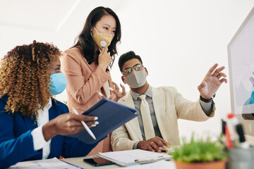 Wall Mural - Business team in protective masks looking at chart on computer screen and discussing sales growth