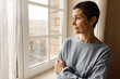 © shurkin_son - Portrait of sad middle aged female with short hairstyle posing by window looking outside with bored pensive facial expression, crossing arms on her chest. People, home and domestic life concept