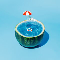 Wall Mural - Watermelon as a pool with crystal clear blue water, parasol, swimming ring, and sunbed on a blue background. Creative summer vacation concept. Tropical fruit diet or idyllic holiday idea.