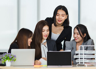 Group of four attractive Asian female office colleagues in formal business suits laughing and working together with computer desktop and laptop on the table in office.