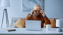 Elderly Man Adjusting Glasses While Working Near Laptop In Living Room.
