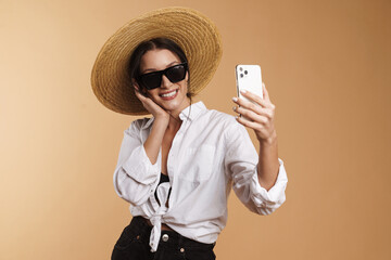 Wall Mural - Young woman in straw hat smiling while taking selfie on cellphone