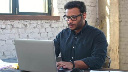 Focused hindu freelancer guy wearing eyeglasses using laptop in contemporary office space, concentrated mixed-race man typing, messaging, indian programmer develops software, looks at computer screen