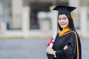 Poster - A young beautiful Asian woman university graduate in graduation gown and mortarboard holds a degree certificate stands in front of the university building after participating in college commencement