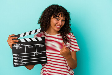 Young mixed race woman holding a clapperboard isolated on blue background pointing with finger at you as if inviting come closer.