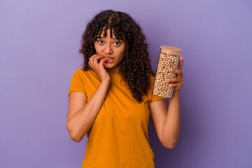 Wall Mural - Young brazilian woman holding a chickpea bottle isolated on purple background biting fingernails, nervous and very anxious.