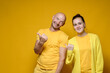 Cute, benevolent man and woman in yellow clothes invite somewhere by pointing their thumb to the side. Copy space. Bright background.