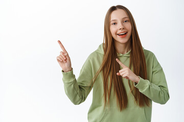 Looh this way. Smiling teen girl demonstrate logo, pointing at banner, showing promo aside, standing in hoodie against white background