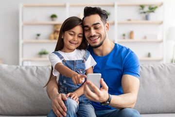 Smiling dad and daughter sitting on couch, using phone