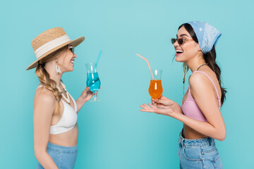 Wall Mural - Cheerful women in sunglasses holding cocktails and talking isolated on blue.