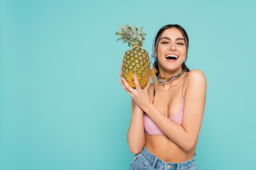 Wall Mural - Positive woman in top holding pineapple and looking at camera isolated on blue.