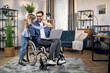 © sofiko14 - Cute daughter embracing her beloved father sitting in wheelchair and using modern laptop. Handicapped man working from home to support his family.