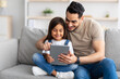 © Prostock-studio - Smiling dad and daughter sitting on couch, using digital tablet