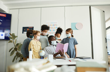 Wall Mural - Portrait of diverse group of children with male teacher wearing masks in school classroom, covid safety measures, copy space