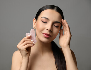 Wall Mural - Beautiful young woman doing facial massage with gua sha tool on grey background