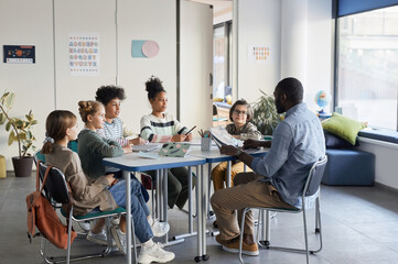 Wall Mural - Full length shot of diverse group of children sitting at table with male teacher in modern school classroom
