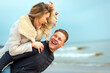 Happy man giving piggyback ride to his woman and laughing at beach. Smiling guy in love carrying on back her girlfriend.