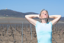 A Blonde Madure Woman Enjoys The Sun In Front Of A Vineyard With Her Arms Raised And Her Eyes Closed. In The Background Is A Tower On Top Of A Hill As Well As Vineyards In Autumn.
