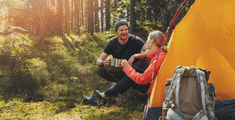 Wall Mural - outdoor camping - young couple resting by a tent with tea cups at forest camp after hiking. active lifestyle