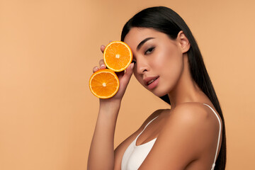 Portrait of Asian girl with shining clean skin of face holding orange halves in white underwear isolated on beige background. Vitamin C cosmetics concept