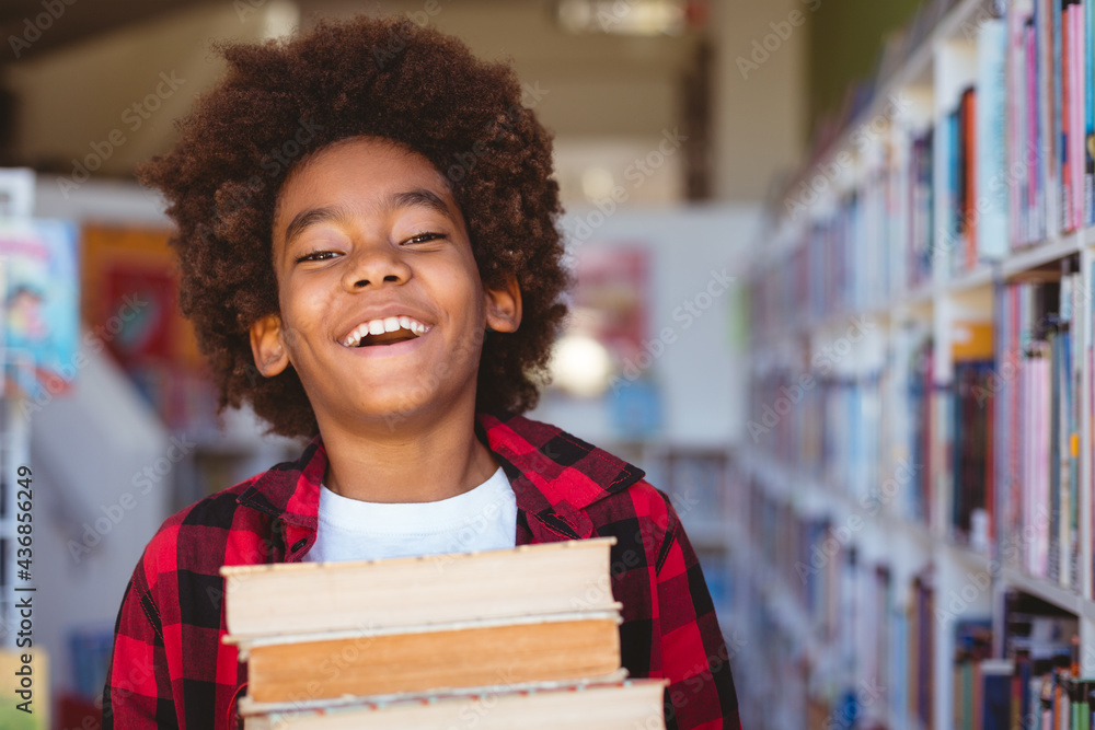 Laughing african american schoolboy carrying stack of books in school library - obrazy, fototapety, plakaty 