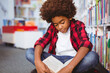 Happy african american schoolboy reading book sitting on floor in school library
