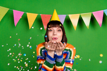 Sticker - Photo of cheerful young happy woman hold hands blow confetti wear cone hat isolated on green color background