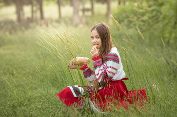 Wall Mural - A girl in a Belarusian folk costume sits in a clearing with homemade bread in her hands. The concept of the traditions of the Slavic people.