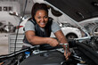African Female Checks an Engine Breakdown. Car Service Employee Woman Fix the Engine Component. Modern Clean Workshop. Black Woman In Overalls Looks At Camera Smiling