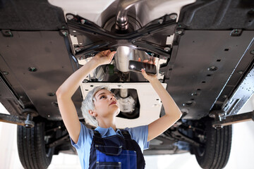 Wall Mural - European Female auto mechanic work in garage, car service technician woman in overalls check and repair customer car at automobile service center, inspecting car under body and suspension system