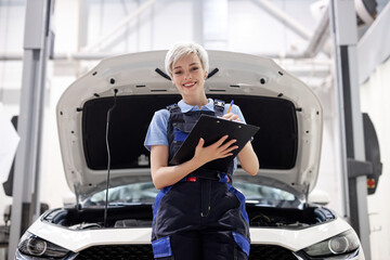 Wall Mural - Portrait of young attractive female in uniform writing information about car in paper tablet, looking at camera and smiling, opened hood of white automobile in the background