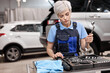 Portrait of young female auto mechanic taking tool instruments for car repairing, having blue rag, box with many various instruments, woman in overalls uniform is ready for work in garage