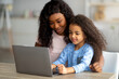 © Prostock-studio - Home schooling during covid concept. Cheerful black mom helping her daughter with online homework on laptop pc