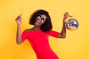 Sticker - Photo of young happy smiling african girl in sunglasses hold disco ball drink champagne isolated on yellow color background