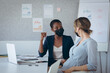 © Wavebreak Media - Two diverse businesswomen wearing face masks, sitting at desk, using laptop, talking