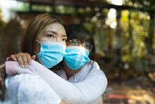 Sad Asian Woman And Her Daughter Embracing Wearing Face Masks And Looking Out Of Window