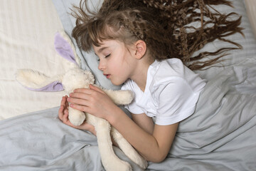 a cute sleeping little girl with a long wavy hair holding a white hare in her hands lying on her sid