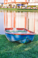 An old battered blue boat with a red rim stands on dry land against a clear lake on a bright sunny day