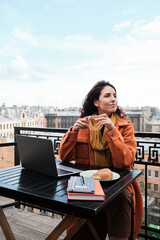 Wall Mural - Young woman sitting at the table with laptop enjoying the cup of tea and looking at view