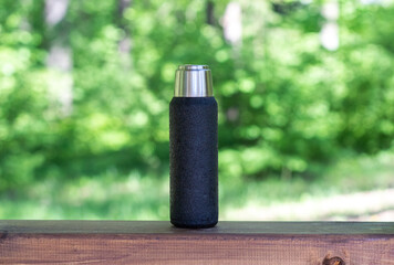 A black thermos with brewed tea or coffee stands on a wooden board with a copyspace. Thermos with hot drink