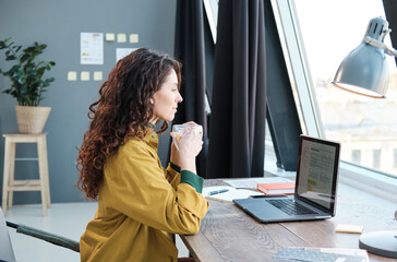 Wall Mural - Young woman drinking coffee while sitting at the table in front of laptop and working at home