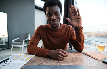 Wall Mural - Portrait of African businessman smiling at camera and waving his hand while sitting at office