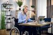 © sofiko14 - Pretty young woman in wheelchair using headset and laptop for video conference at home. Handicapped business lady leading working meeting online.
